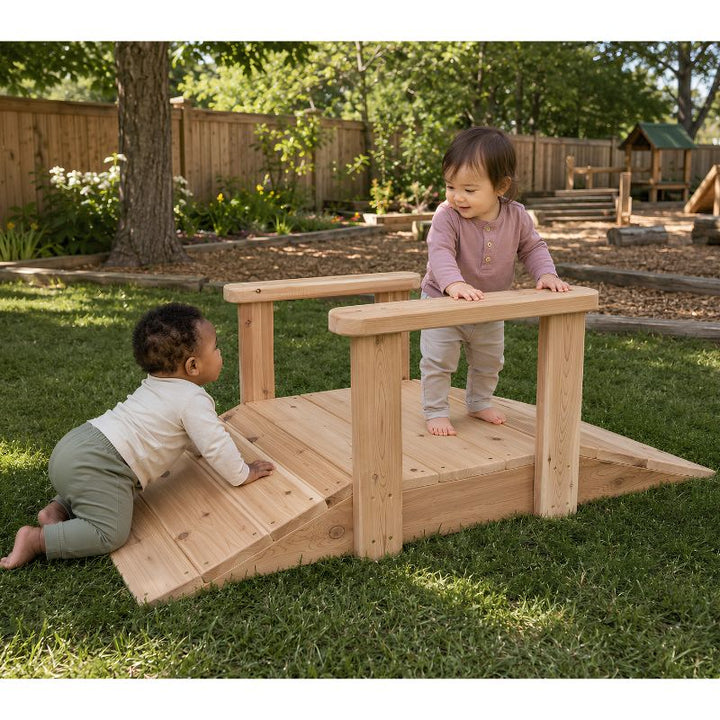 An infant crawling up the wooden ONP Infant Ramp outdoors while a toddler stands at the top holding the handrail