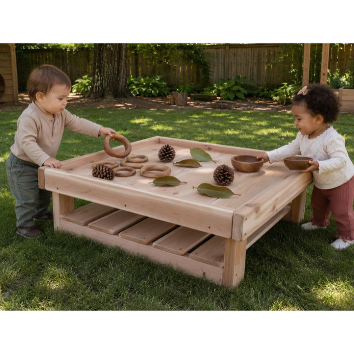 Two toddlers playing at the low ONP Infant Square Table outdoors, sorting wooden rings, bowls, pinecones and leaves on the tray surface