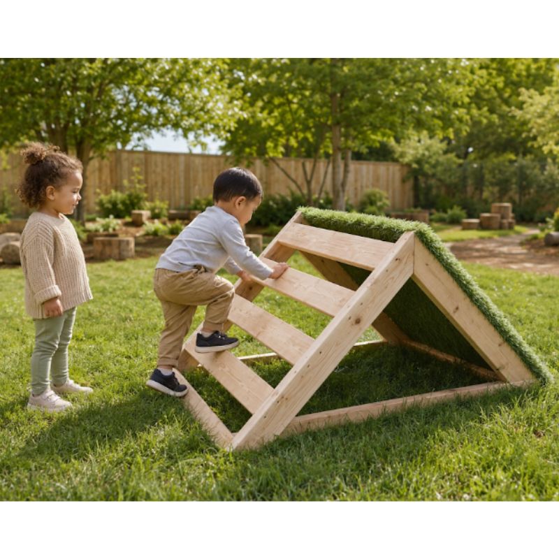 A toddler boy climbing the wooden ladder side of the ONP Mini-Climber outdoors, with a turf-covered ramp on the opposite side