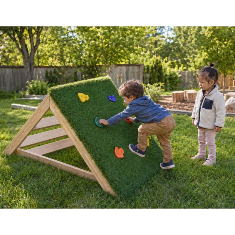 A toddler boy climbing the turf-covered side of the ONP Mini-Climber using coloured grip holds, while a girl watches nearby