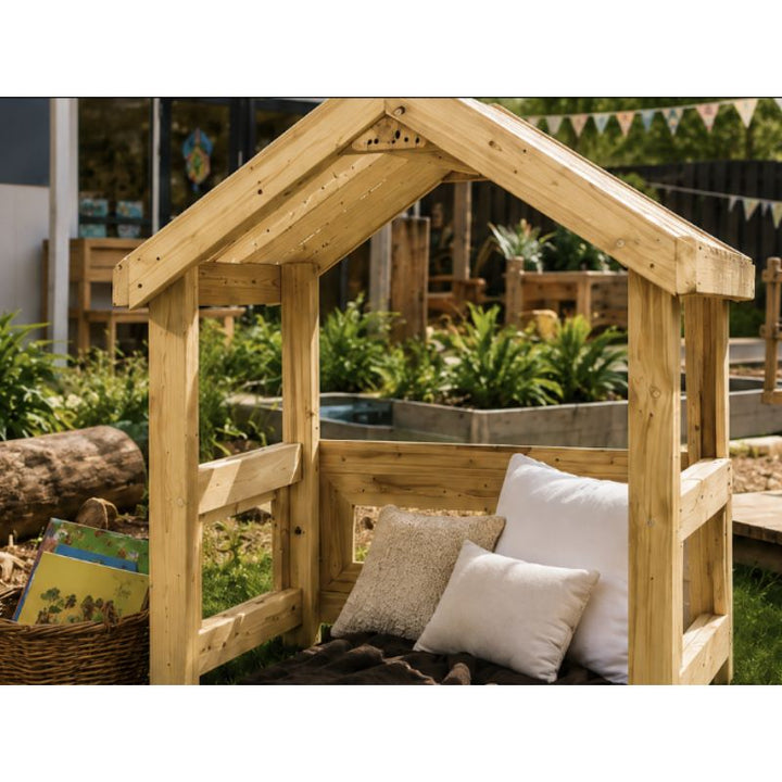 Close-up of the ONP Outdoor Baby Den outdoors, showing the peaked wooden frame with cushions, a blanket and a basket of books