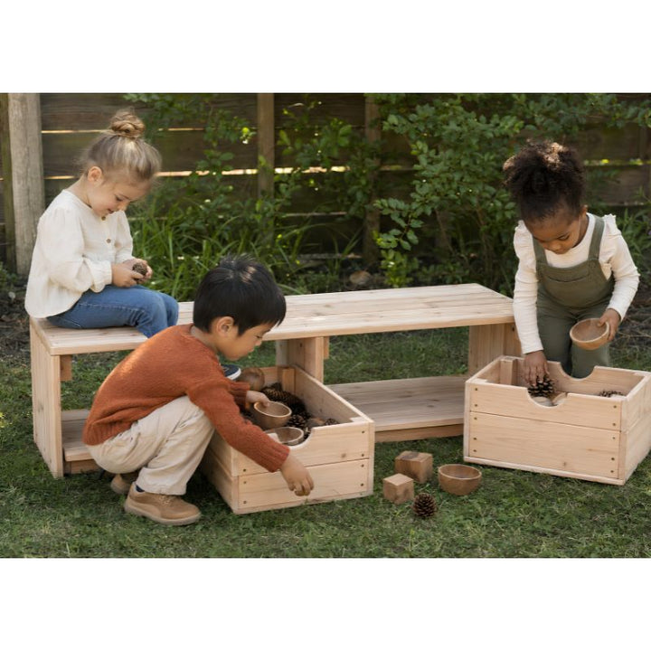Three children playing at the ONP Outdoor Bench and Storage outdoors, sorting pinecones and blocks into the pull-out crates beneath the bench