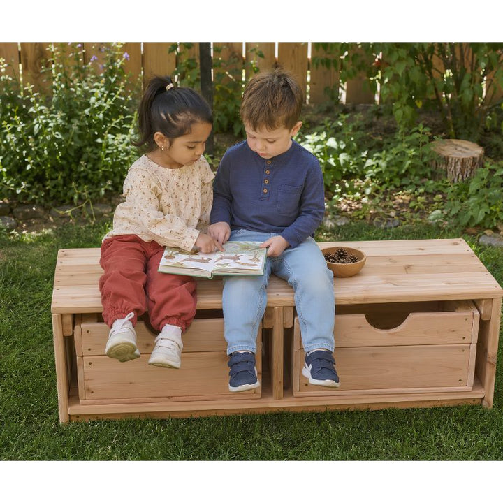 Two children reading a book while seated on the ONP Outdoor Bench and Storage outdoors, with wooden crates tucked into the base