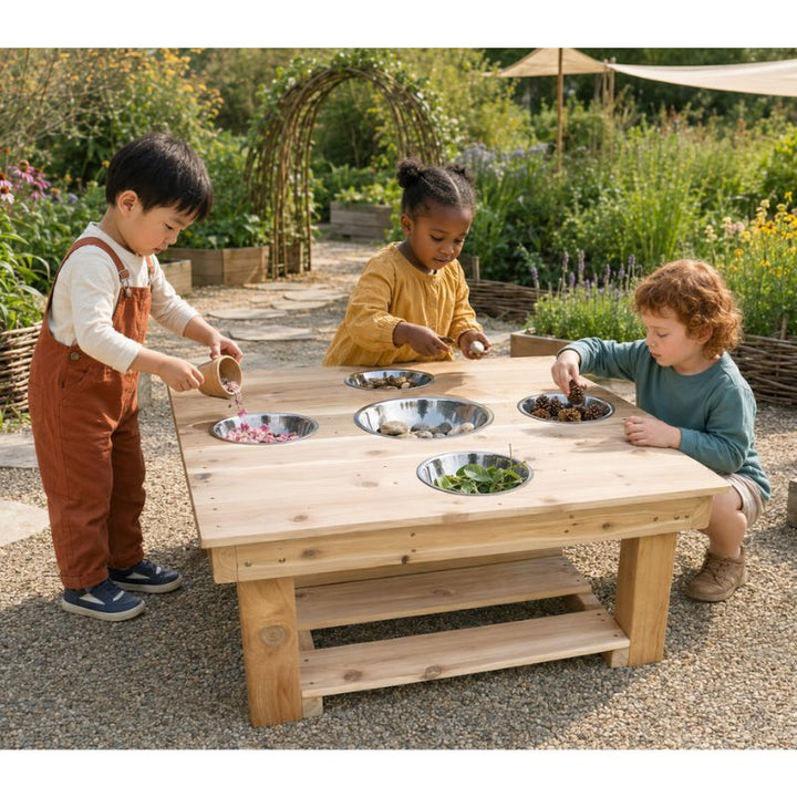 Three children gathered around the ONP Outdoor Messy Activity Table in a garden, filling the five stainless steel bowls with petals, stones, pinecones and leaves