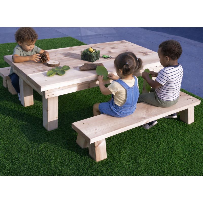 Three children seated at a wooden picnic table and benches outdoors, exploring leaves, a pinecone and a wood slice with toy bugs