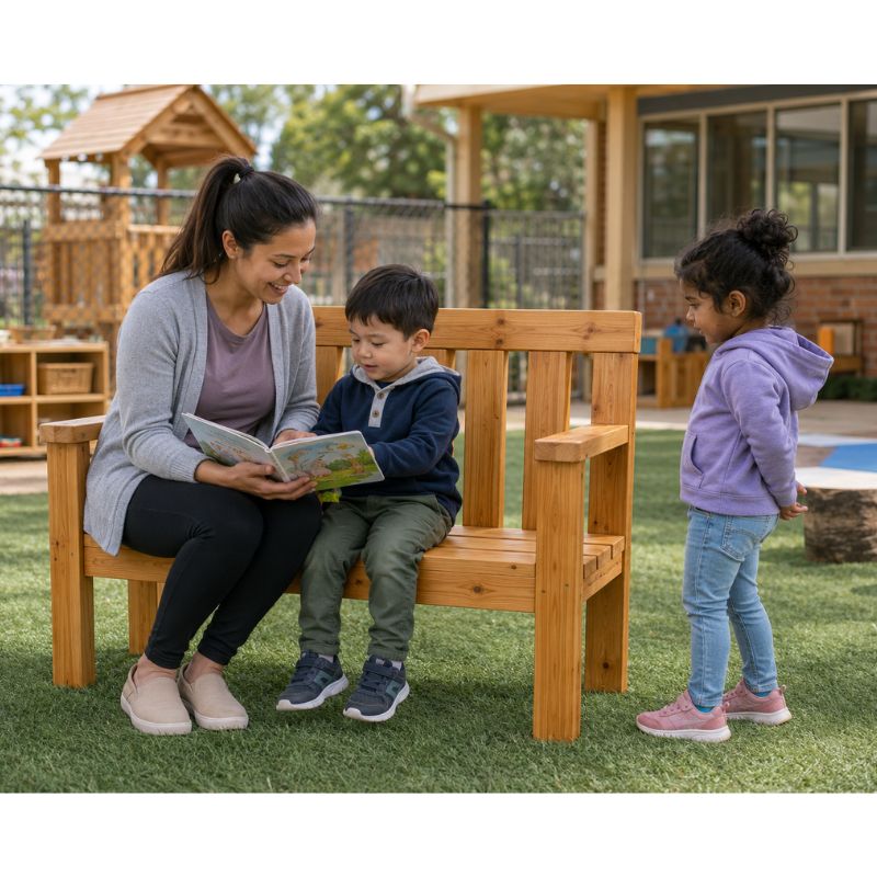 A woman reading a book with two children seated on the ONP Preschool Buddy Bench outdoors, with a playground visible in the background