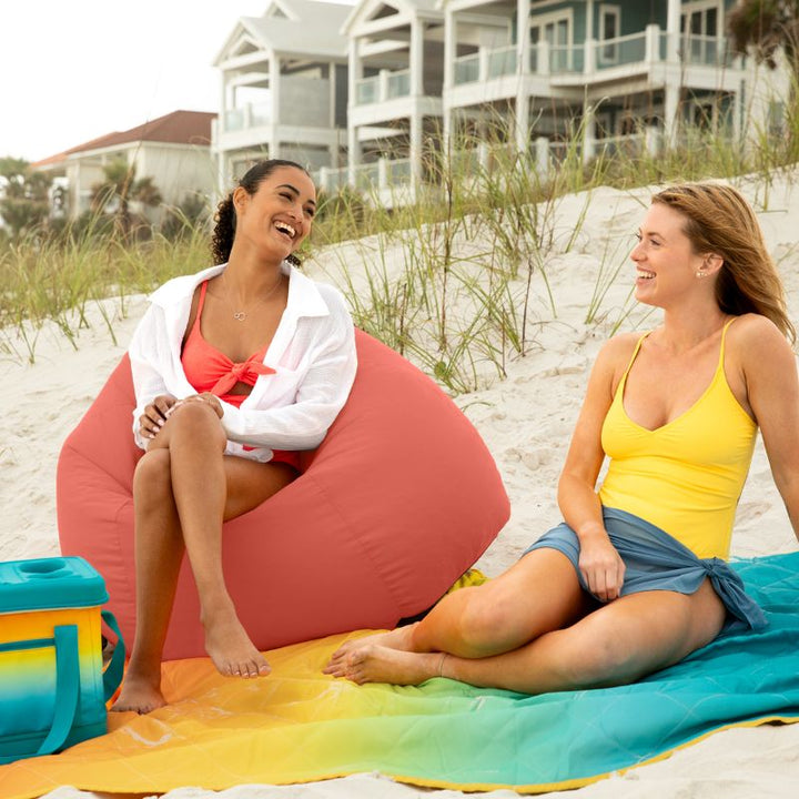 Two women laughing together, one seated in a Flamingo Jaxx Kiss outdoor bean bag chair on the beach