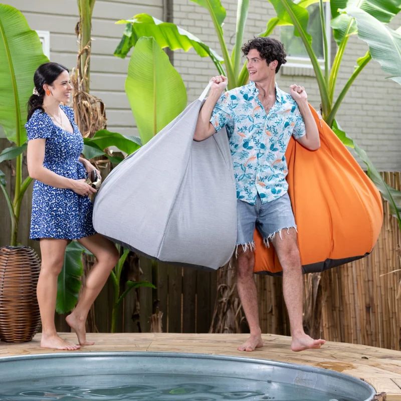 Man and woman carrying Granite and Tangerine Jaxx Kiss outdoor bean bag chairs beside a pool