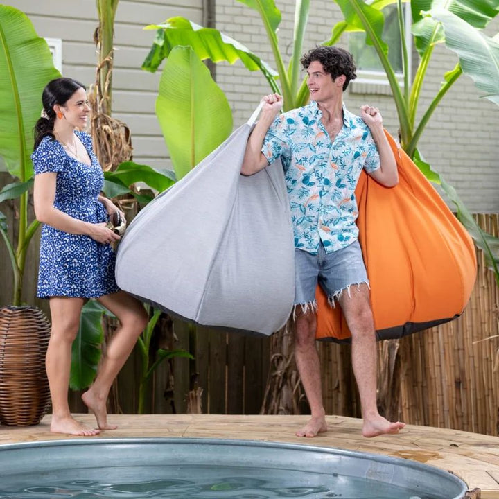 Man and woman carrying Granite and Tangerine Jaxx Kiss outdoor bean bag chairs beside a pool