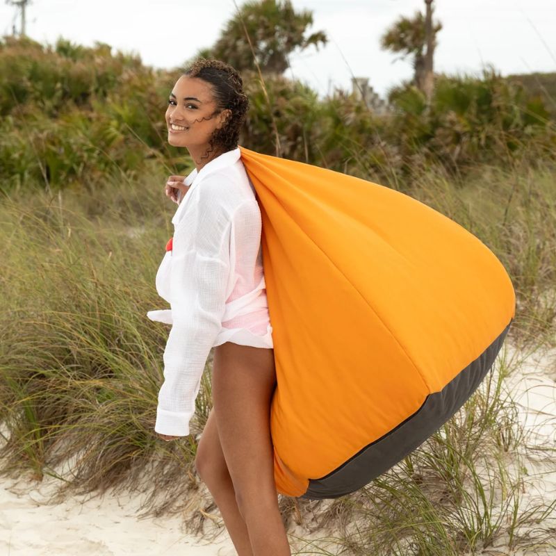 Woman carrying a Tangerine Jaxx Kiss outdoor bean bag chair by its reinforced handle on the beach