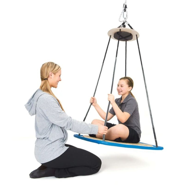 Adult kneeling beside a smiling child on the Southpaw Platform Swing with carpet, guiding tilting vestibular movement during a sensory integration session