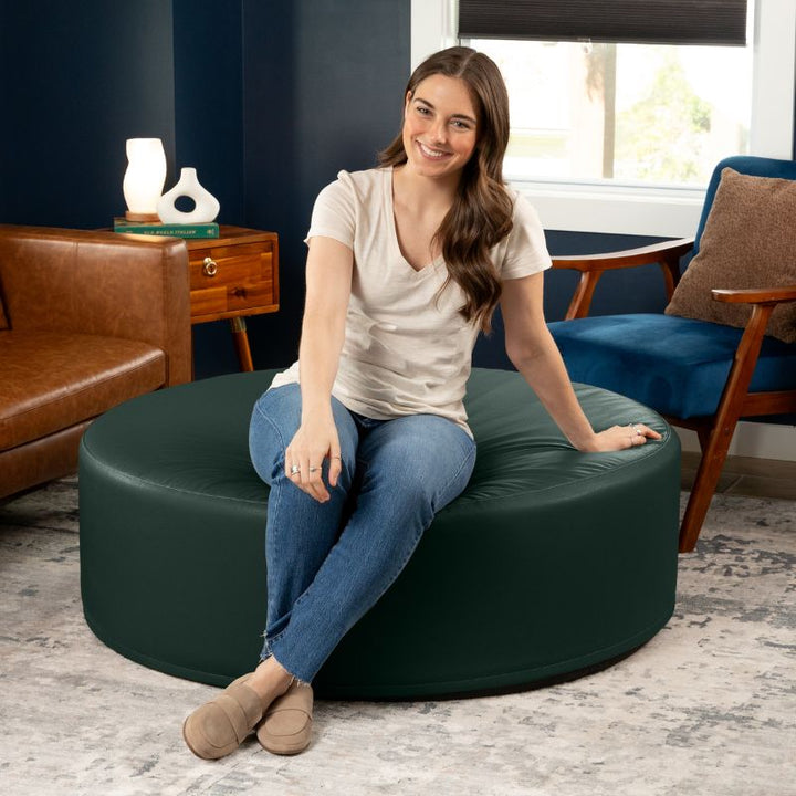 Woman seated on the Jaxx Candler round ottoman in dark green, showing the scale of this flexible foam seating