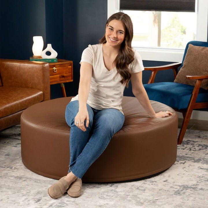 Woman seated on the Jaxx Candler round ottoman in warm brown, showing the scale of this flexible foam seating