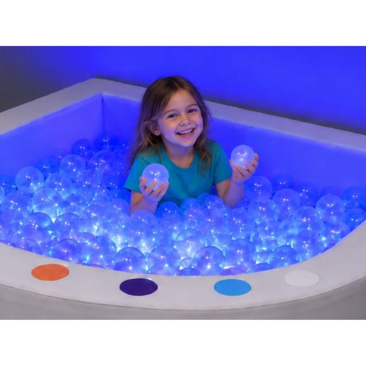 Smiling child holding clear balls lit blue in the Experia Interactive LED Ball Pool, a sensory ball pit for play and calm