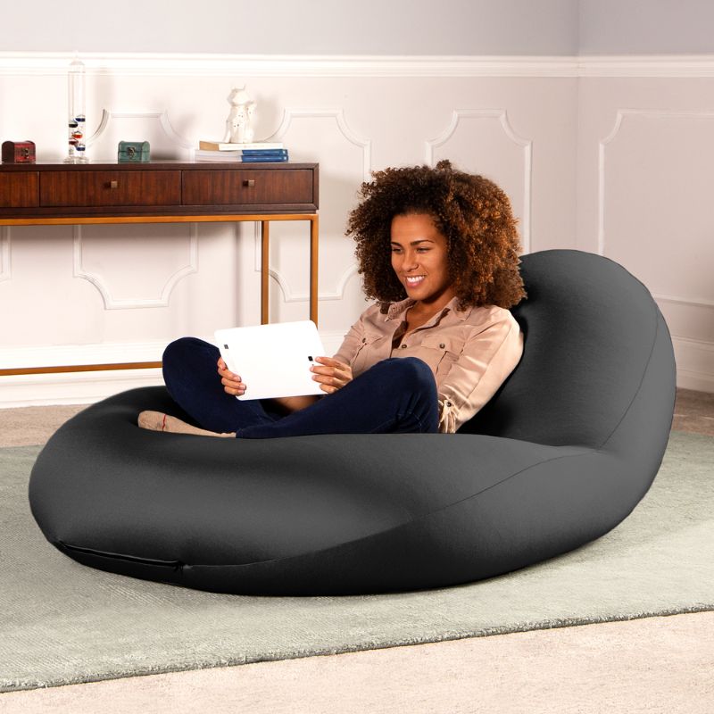 A woman sits with a tablet in the black Jaxx Nimbus sensory bean bag chair in a living room, showing the stretchy cover as home seating