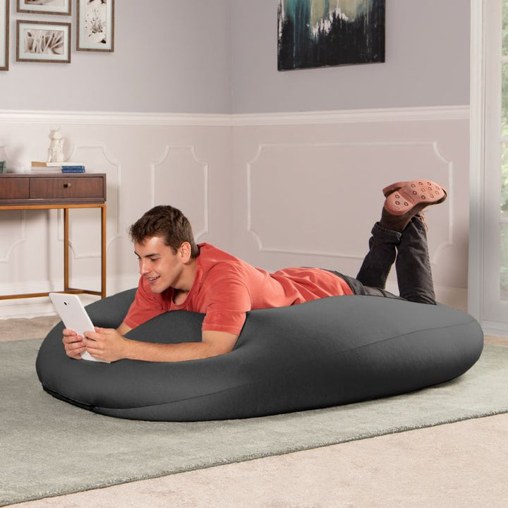 A man lies on the black Jaxx Nimbus sensory bean bag chair using a tablet, showing the flat, flexible seating position
