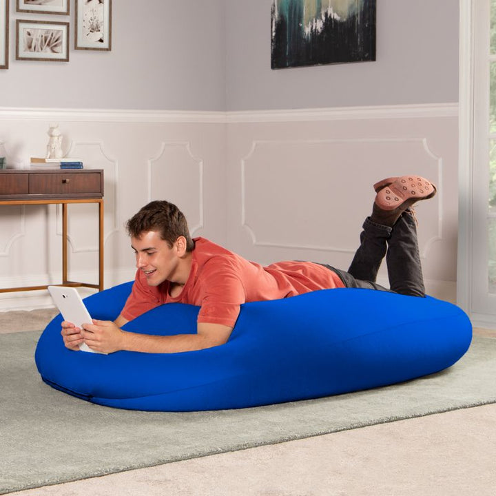 A man lies on the blue Jaxx Nimbus sensory bean bag chair using a tablet, showing the stretchy spandex cover in a lounging position