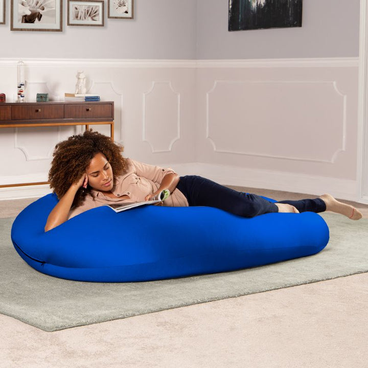 A woman reads a book lying across the blue Jaxx Nimbus sensory bean bag chair, showing the stretchy spandex cover in a lounging position