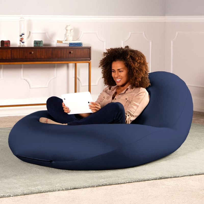 A woman sits with a tablet in the navy Jaxx Nimbus sensory bean bag chair in a living room, showing flexible sensory seating for home use