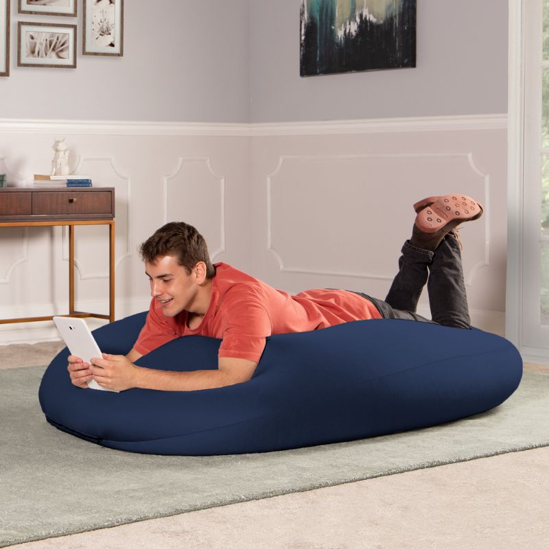 A man lies on the navy Jaxx Nimbus sensory bean bag chair using a tablet, showing flexible sensory seating in a lounging position