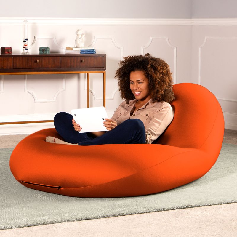A woman sits with a tablet in the orange Jaxx Nimbus sensory bean bag chair in a living room, showing flexible seating for home use