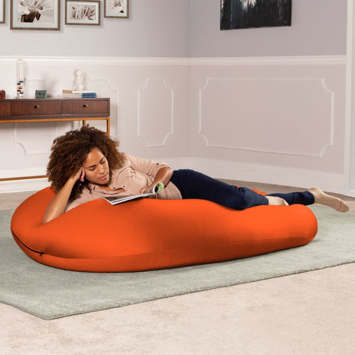 A woman reads a book lying across the orange Jaxx Nimbus sensory bean bag chair, showing the stretchy spandex cover in a lounging position