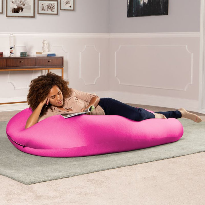 A woman reads a book lying across the pink Jaxx Nimbus sensory bean bag chair, showing the flat, flexible seating position