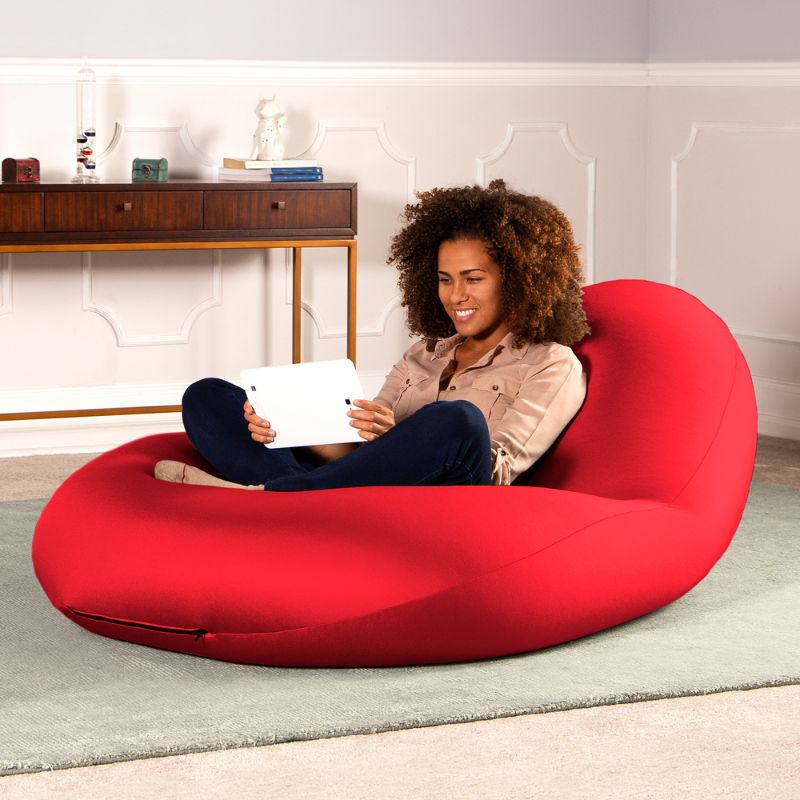 A woman sits with a tablet in the red Jaxx Nimbus sensory bean bag chair in a living room, showing the stretchy cover as home seating