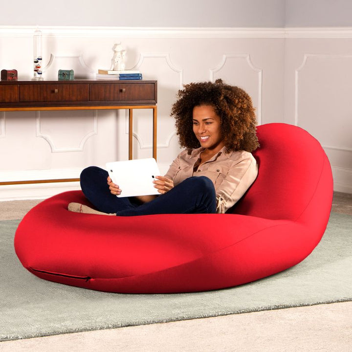 A woman sits with a tablet in the red Jaxx Nimbus sensory bean bag chair in a living room, showing the stretchy cover as home seating