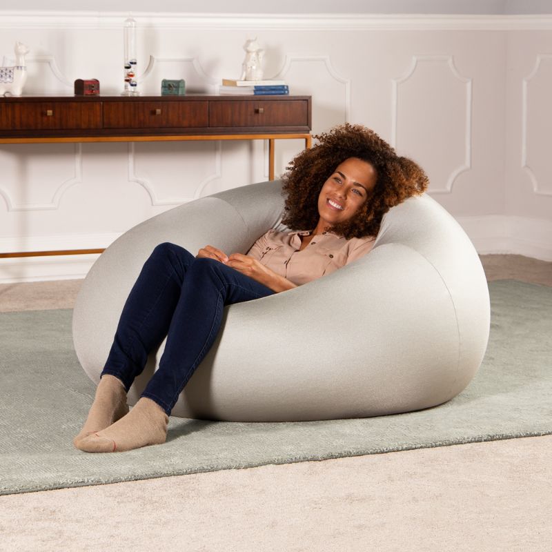 A woman relaxes in the silver Jaxx Nimbus sensory bean bag chair in a living room, showing the stretchy cover as home seating