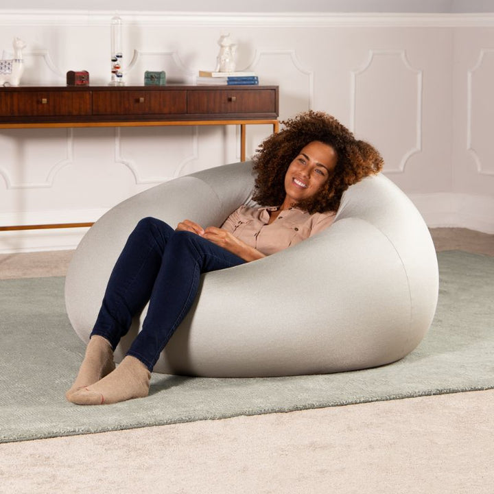 A woman relaxes in the silver Jaxx Nimbus sensory bean bag chair in a living room, showing the stretchy cover as home seating