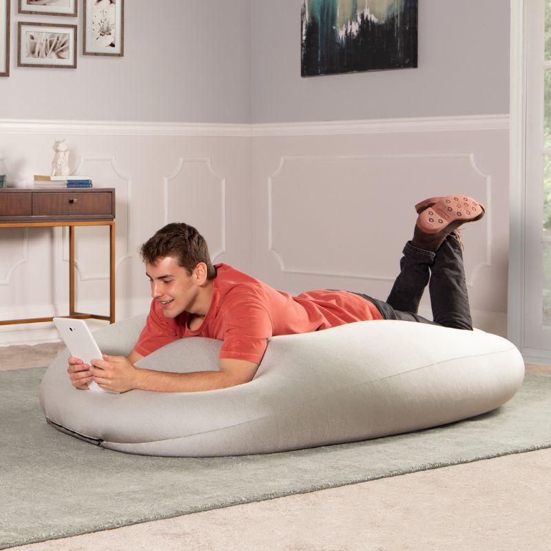 A man lies on the light gray Jaxx Nimbus sensory bean bag chair using a tablet, showing the flat, flexible seating position