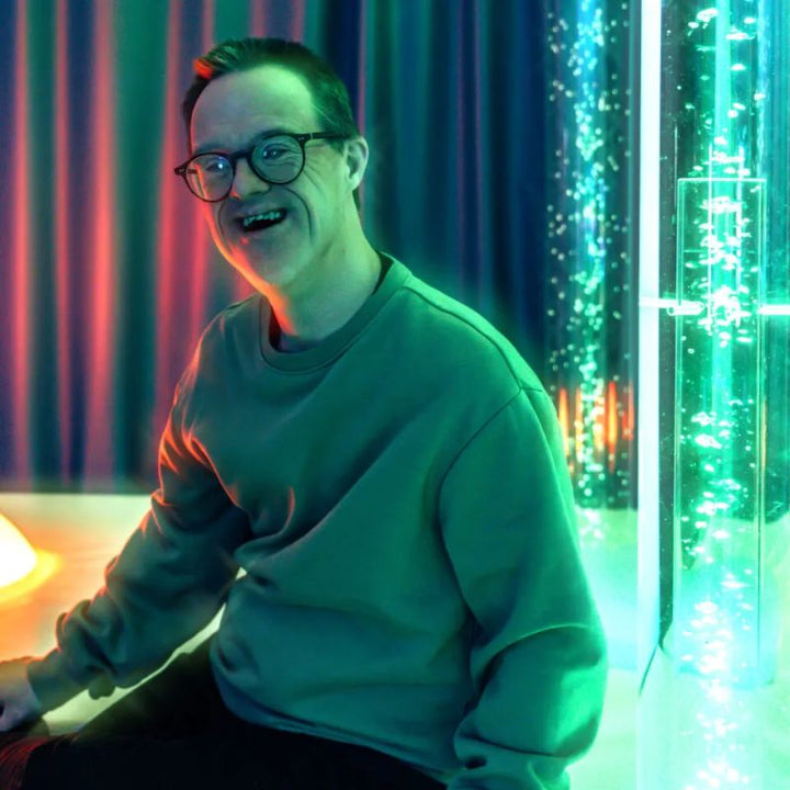 Smiling adult relaxing beside the TFH Giant Water Bubble Tube 8 inch wide in a multi-sensory room, a giant bubble tube for therapy spaces of all kinds