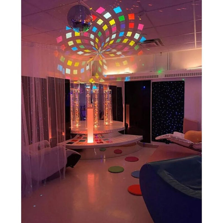 Cluster of TFH Interactive LED Bubble Tubes on a raised platform in a multi-sensory room with mirror ball lighting, soft seating and floor spots