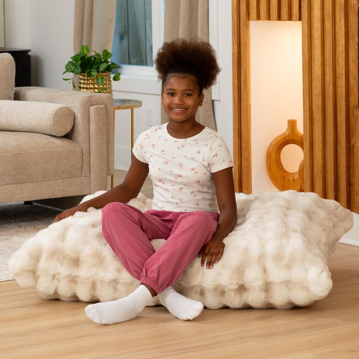 Girl seated cross-legged on the CrashPuff by Raven-Symoné faux fur floor lounger in Phantom Cream