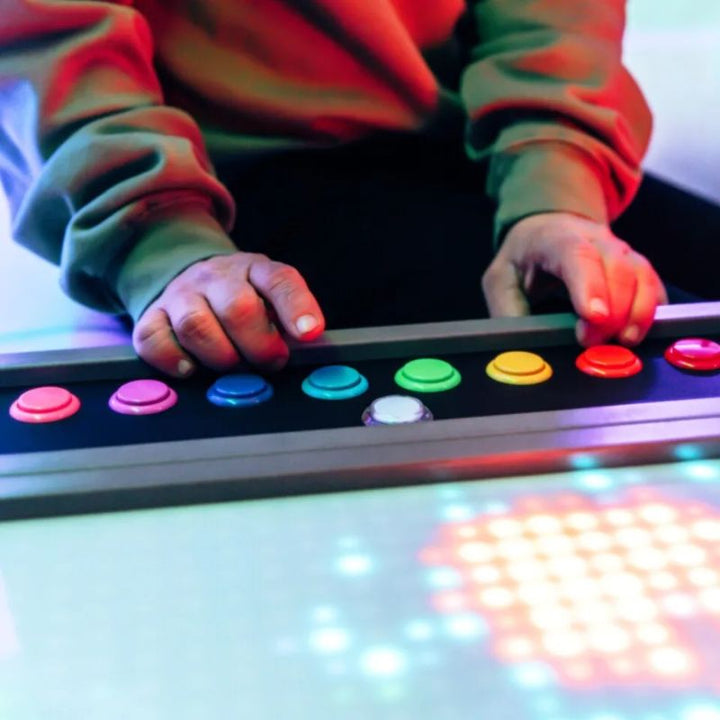 Hands resting on the rainbow colour buttons of the TFH Pixels Pro Illumination Panel, an interactive sensory light panel for cause-and-effect play