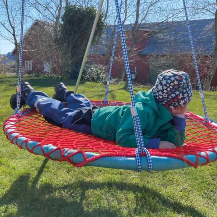 A child lying back on the TFH Nest Swing outdoors on a sunny day, resting in the basket seat of this sensory swing