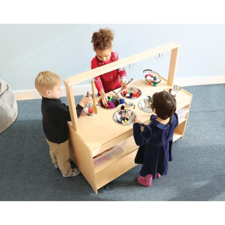Three children using tongs to sort colorful pom-poms in the stainless steel bowls of the Whitney Brothers Mobile Sensory Play Kitchen during group sensory table play