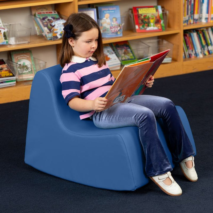 A child reading in the blue Jaxx Midtown Rocker, a sensory rocking chair for kids in a school library