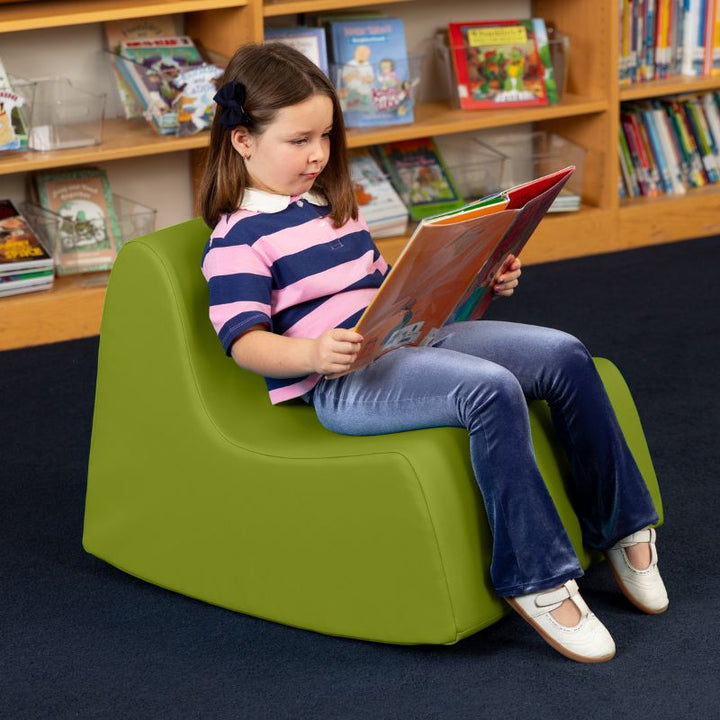 A child reading in the green Jaxx Midtown Rocker, a sensory rocking chair for kids in a school library