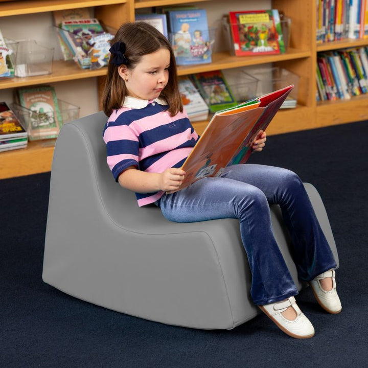 A child reading in the grey Jaxx Midtown Rocker, a sensory rocking chair for kids in a school library
