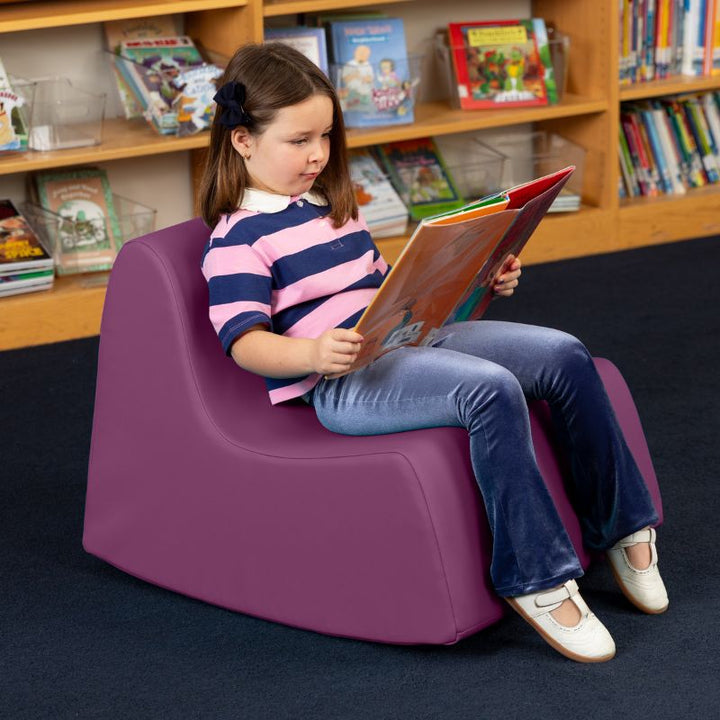 A child reading in the purple Jaxx Midtown Rocker, a sensory rocking chair for kids in a school library