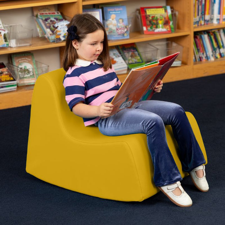 A child reading in the yellow Jaxx Midtown Rocker, a sensory rocking chair for kids in a school library