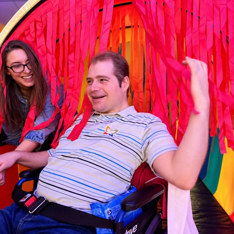 Smiling adult using a wheelchair reaching through the red ribbons of the TFH Giant Tickle Me Tunnel, a tactile sensory tunnel for all ages and abilities