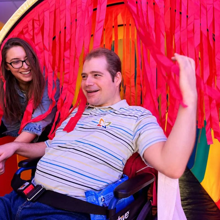 Smiling adult using a wheelchair reaching through the red ribbons of the TFH Giant Tickle Me Tunnel, a tactile sensory tunnel for all ages and abilities
