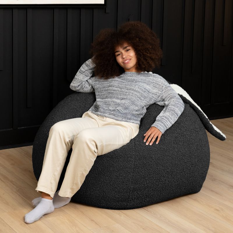 an adult reclines in the black Raven-Symoné HopSeat sherpa bean bag chair, sinking into its deep-pressure comfort