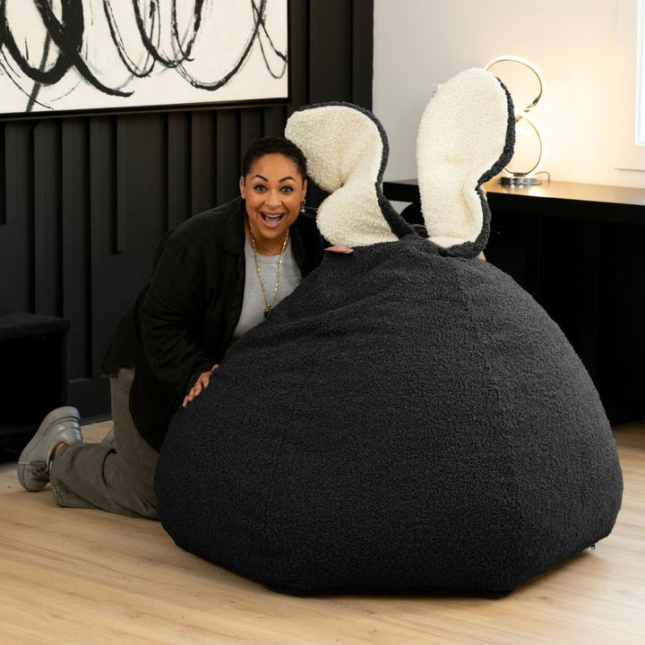 n adult playfully peeks over the black Raven-Symoné HopSeat sherpa bean bag chair, showing its tall bunny-ear design