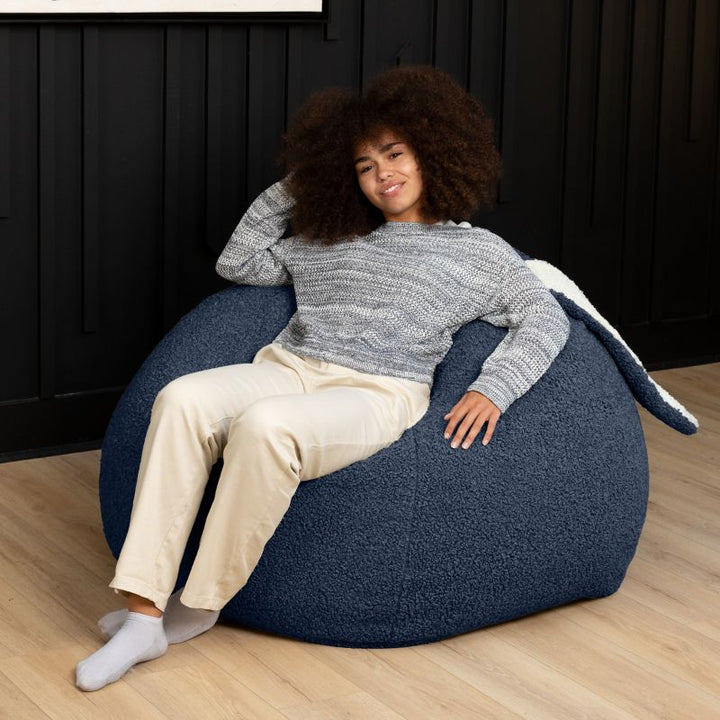 an adult reclines in the blue Raven-Symoné HopSeat sherpa bean bag chair, sinking into its deep-pressure comfort
