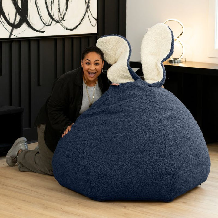 an adult playfully peeks over the blue Raven-Symoné HopSeat sherpa bean bag chair, showing its tall bunny-ear design