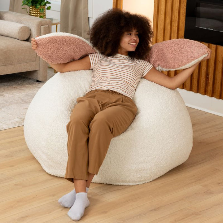 an adult holds up the soft bunny ears of the Cloud Raven-Symoné HopSeat sherpa bean bag chair in a bright living room